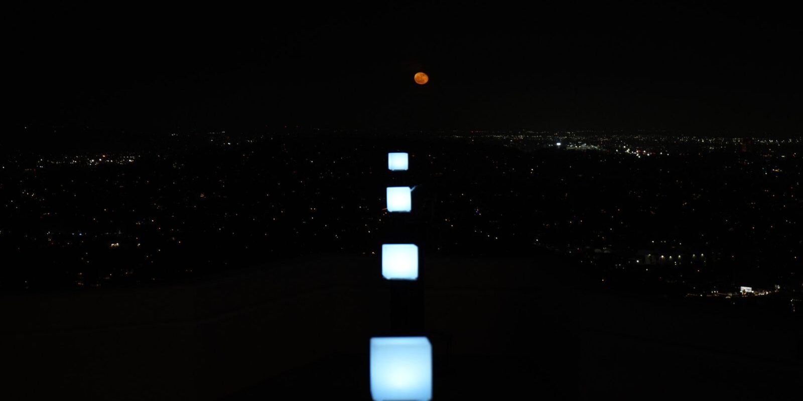 Square white lights in line with the path of a coppery red moon rise.