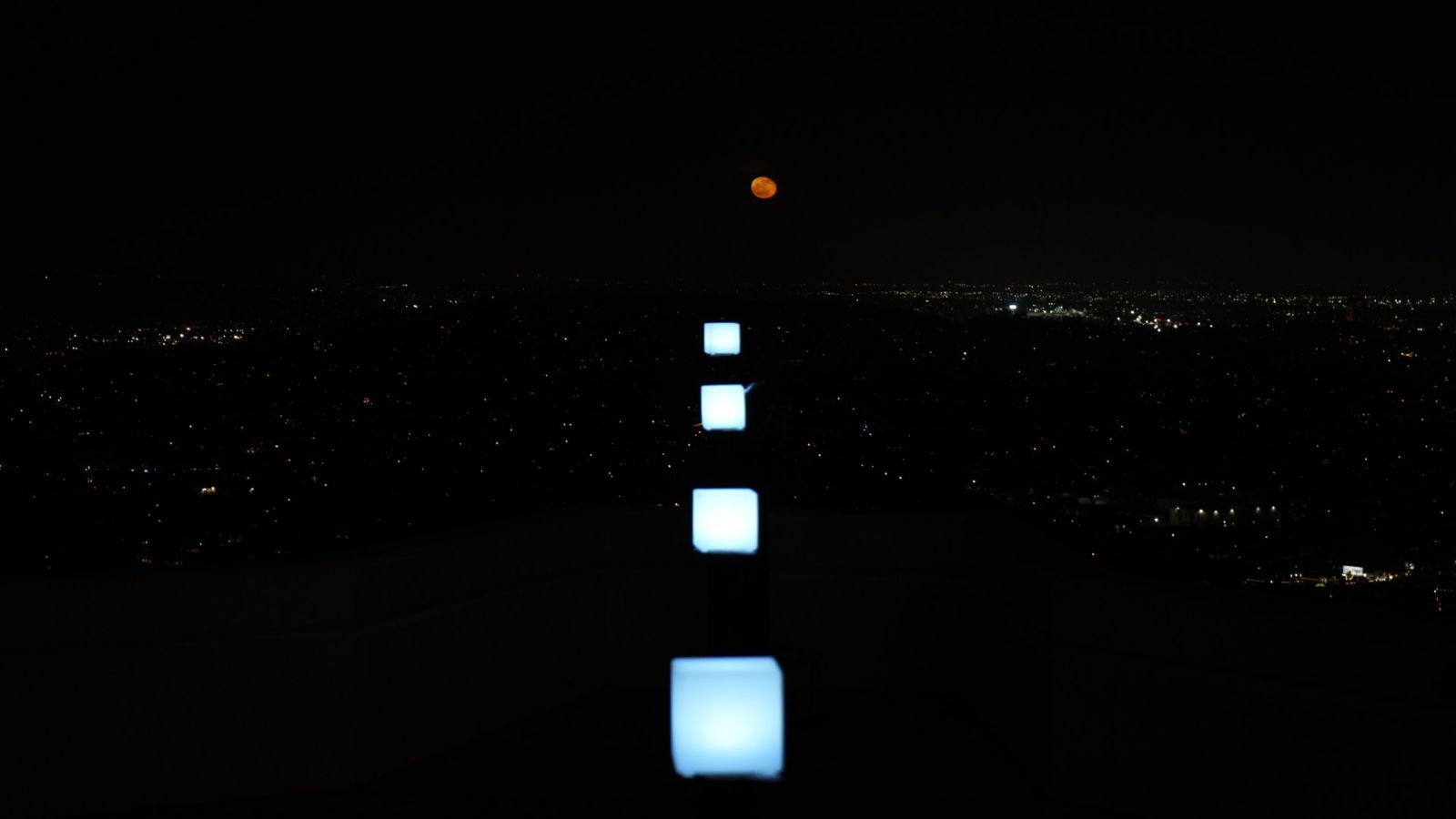 Square white lights in line with the path of a coppery red moon rise.