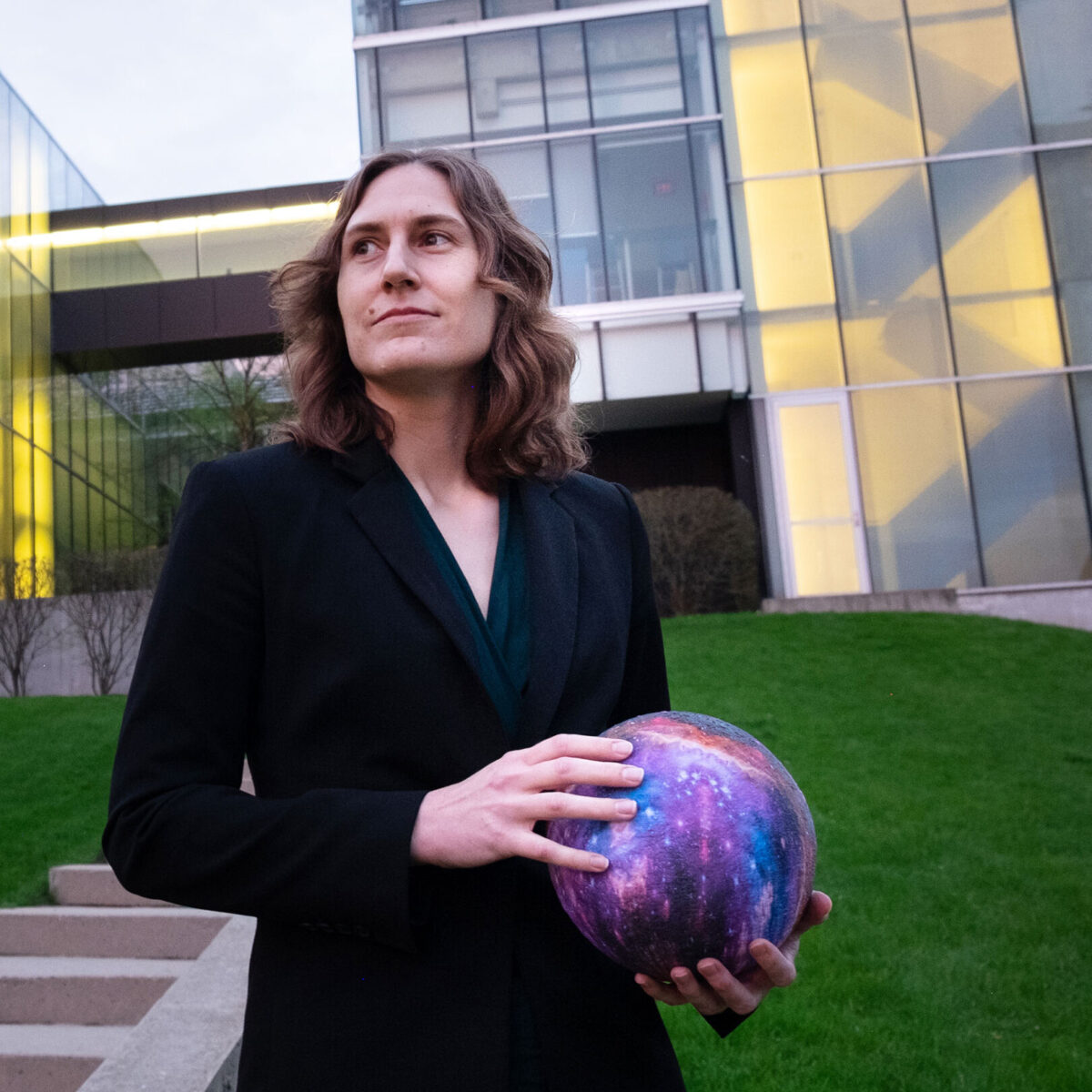 Guest speaker Dr. Katie Mack holds a globe in front of a modern building and gazes confidently into the distance.