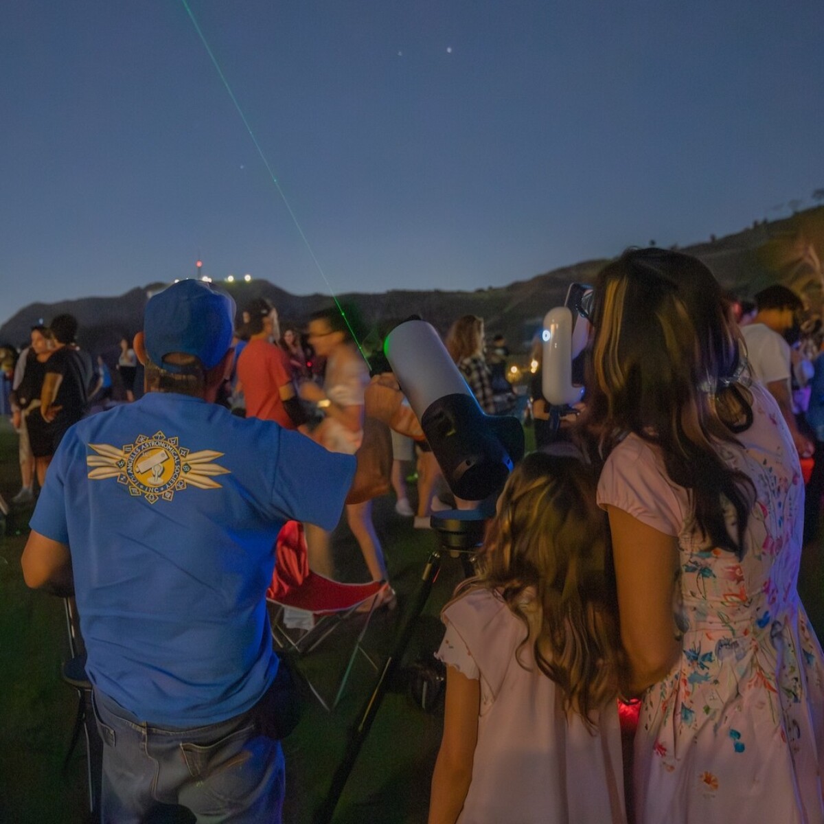 A telescope operator pointing out an object in the night sky while two patrons look up.