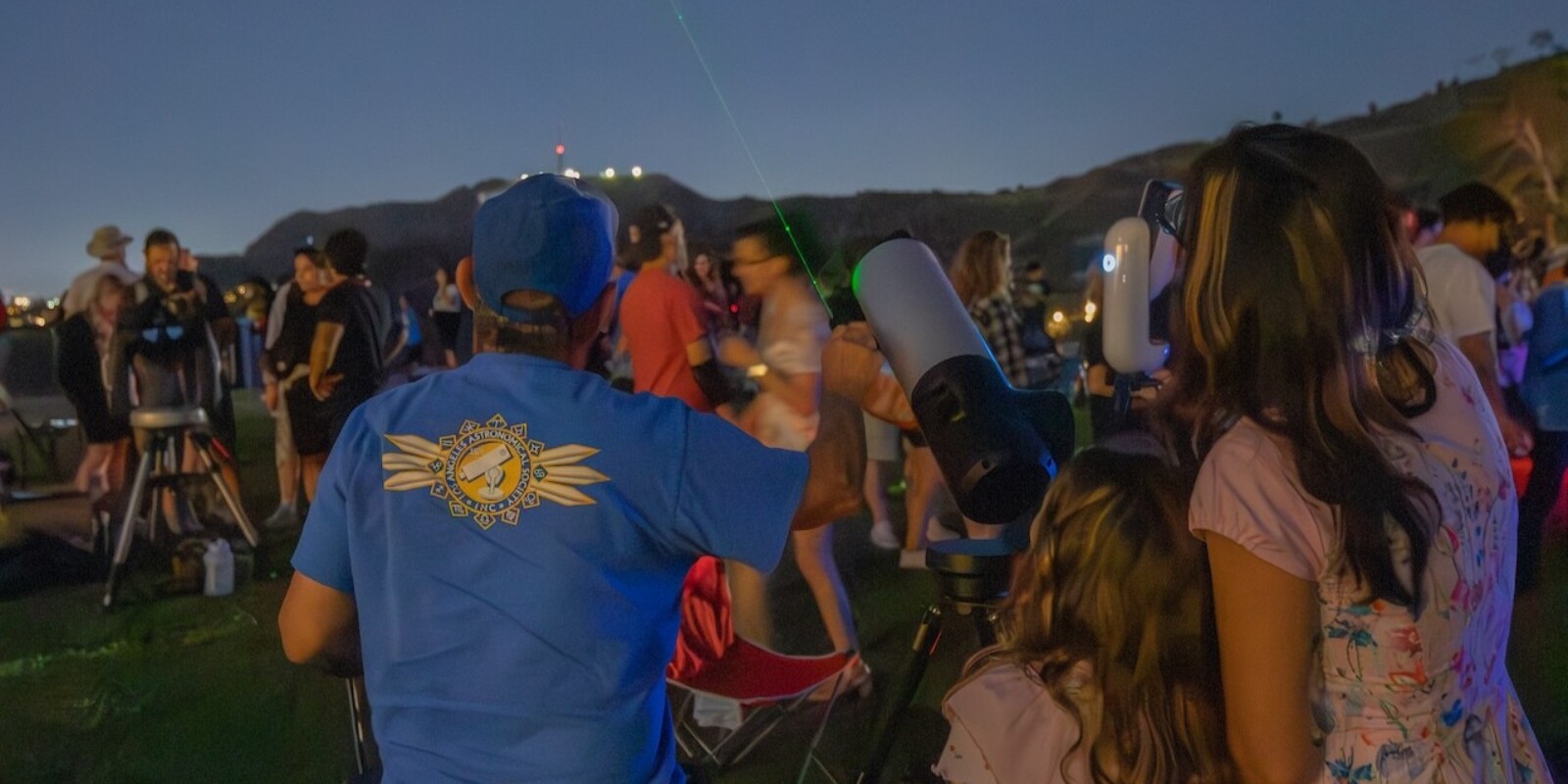 A telescope operator pointing out an object in the night sky while two patrons look up.