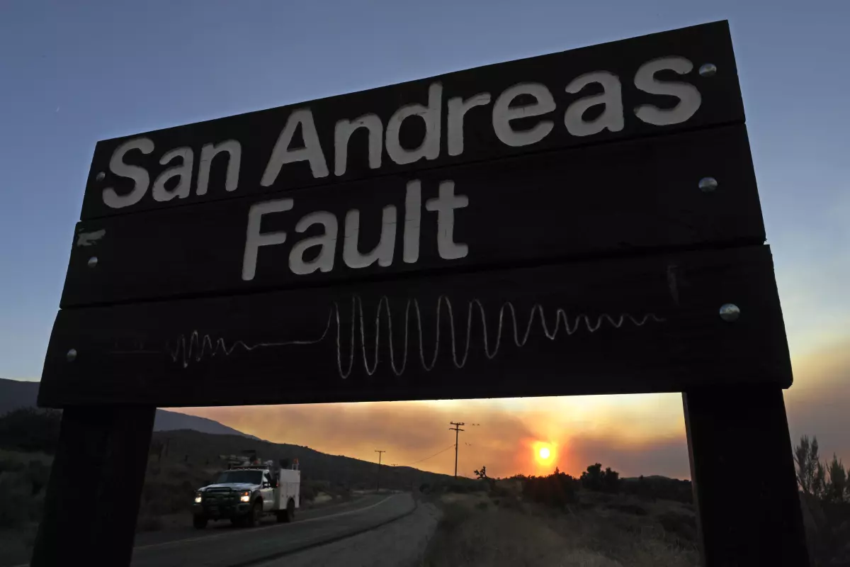 A wooden sign reads "San Andreas Fault" with a sine wave illustration. A road with a truck and a sunset are in the background.