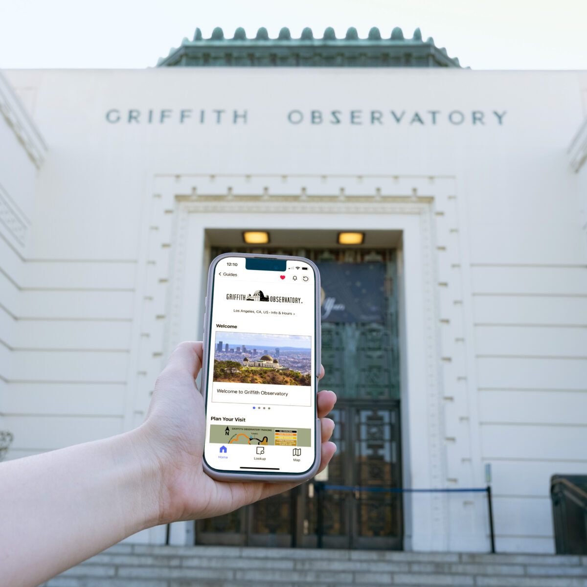 A woman's hand stretches into the frame with a smartphone that displays the Bloomberg Connects app. Griffith Observatory sits in the background.