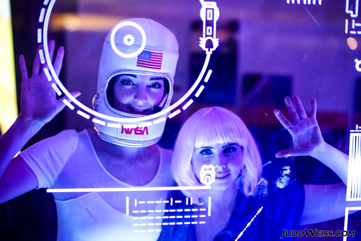 Two women press their hands to a glass screen with sci-fi vinyl decals in a room lit in purple. One woman wears a plush astronaut helmet costume piece, and the other wears a platinum blonde wig.